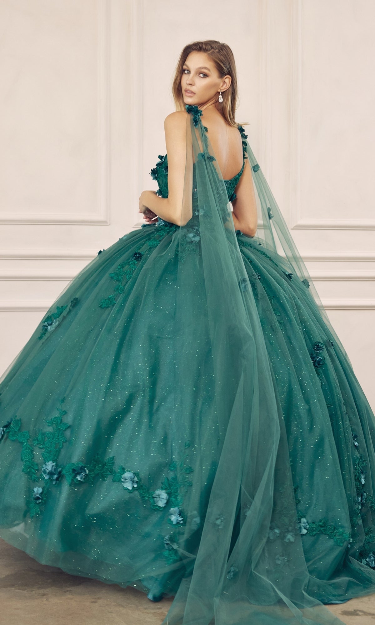 Woman wearing the Juliet 1436 Glitter Quinceañera Dress with Cape, featuring floral details and long tulle accents, poses gracefully in front of a white paneled wall.